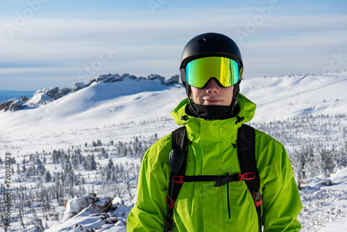 Close-up portrait of a skier or snowboarder in sports equipment, snowy mountains background at ski resort. Bright acid green outfit: warm suit jacket, goggles, black sport helmet, backpack straps photo