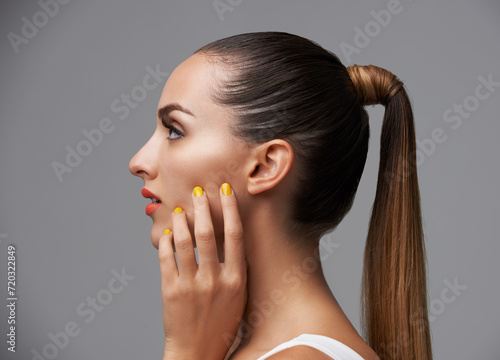 Manicure, face and profile of woman in makeup, beauty and cosmetics with nails and hair care on grey background. Hand, yellow nail polish and lipstick with skin, glamour and shiny ponytail in studio