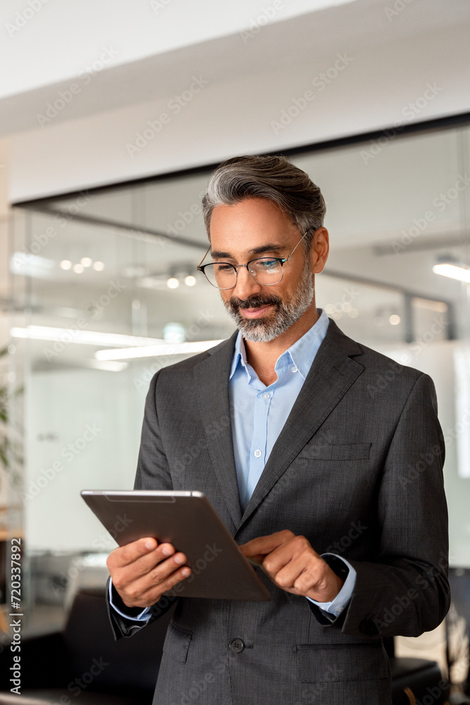 Vertical portrait of smiling Hispanic mature adult professional business man, happy Indian 40s 50s businessman CEO holding digital tablet using fintech tab application standing inside company office. Vertical portrait of smiling Hispanic mature adult professional business man, happy Indian 40s 50s businessman CEO holding digital tablet using fintech tab application standing inside company office.