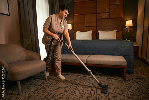 Uniformed maid is cleaning guest suite with vacuum cleaner