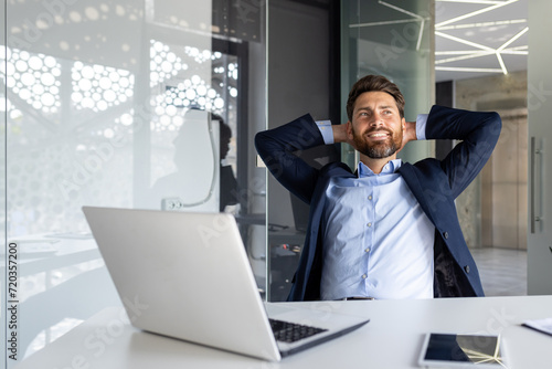 Happy and smiling young businessman man sitting in the office at the desk and resting and satisfied with the work with his hands behind his head photo