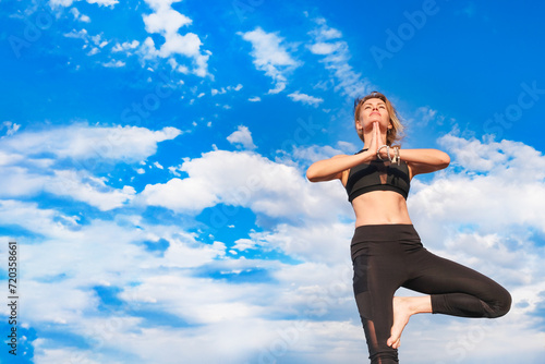 Slender blonde girl doing yoga outside against a blue sky on a sunny day.