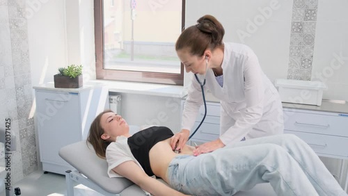 A female doctor with a stethoscope examines the abdomen of a young patient lying on a bed in the emergency department of a hospital