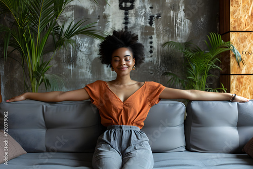 a young afro woman sitting on a gray couch and stretches her arms out © Yi_Studio