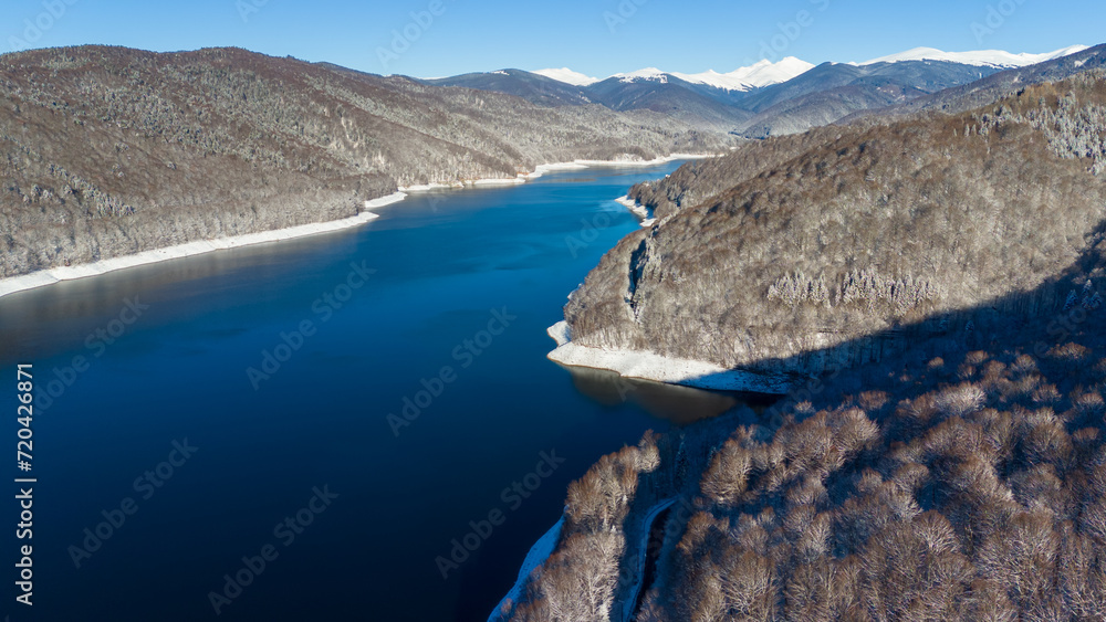Mountain lake surrounded by forest. Mountain peaks covered with snow. Lake Vidraru dam in Romanian mountains Fagaras. Aerial photography.