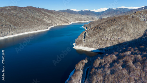 Obraz na płótnie Mountain lake surrounded by forest. Mountain peaks covered with snow. Lake Vidraru dam in Romanian mountains Fagaras. Aerial photography. w salonie