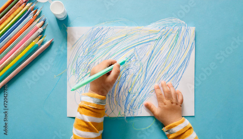 Baby hand holding marker and drawing colorful scratches lines on white paper. Blue table background. Pastel color. Closeup. Toddler development. Learning painting. Point of view shot. Top down view