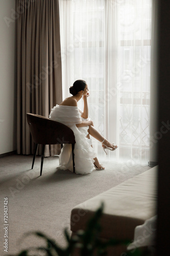 Morning and getting ready for a luxurious and beautiful bride in a hotel in a luxury room in pajamas and lingerie, putting on shoes and a chic white wedding dress