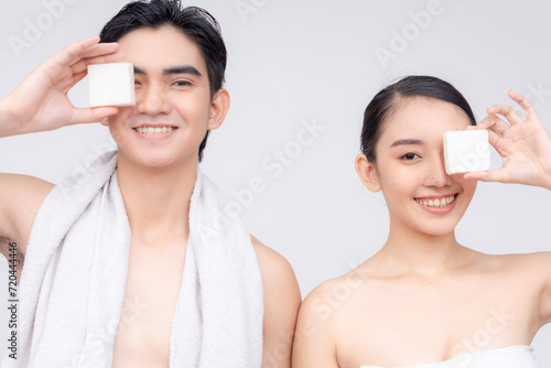 A young asian couple in their early 20s wrapped in white towels poses with a white collagen soap. Isolated on a white backdrop.