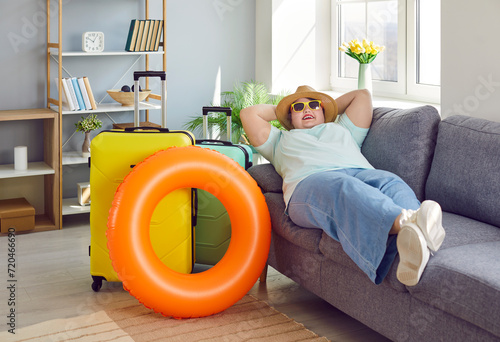 Happy plus size woman relaxing on sofa with suitcase and inflatable ring in living room at home. Happy overweight, obese woman in casual clothes, hat and sunglasses going on summer vacation