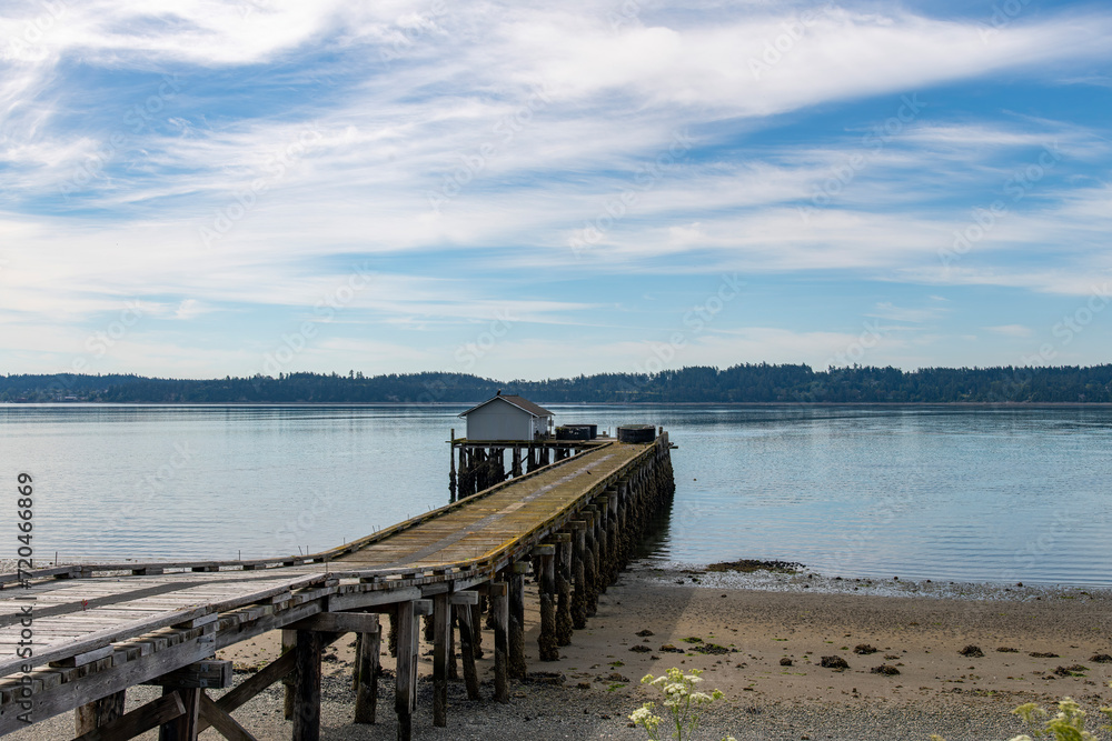 Panoramic view over the water of Penn Cove near Coupeville, WA, USA with a wooden fishing dock and building extended in the water against a blue sky with white feather clouds 