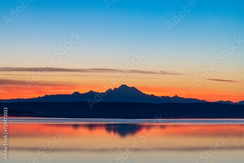 Colorful panoramic view early summer morning sunrise from Coupeville, WA, USA over Skagit Bay towards stratovolcano Mt Baker in Cascades Mountain range in silhouette and reflection in tranquil water