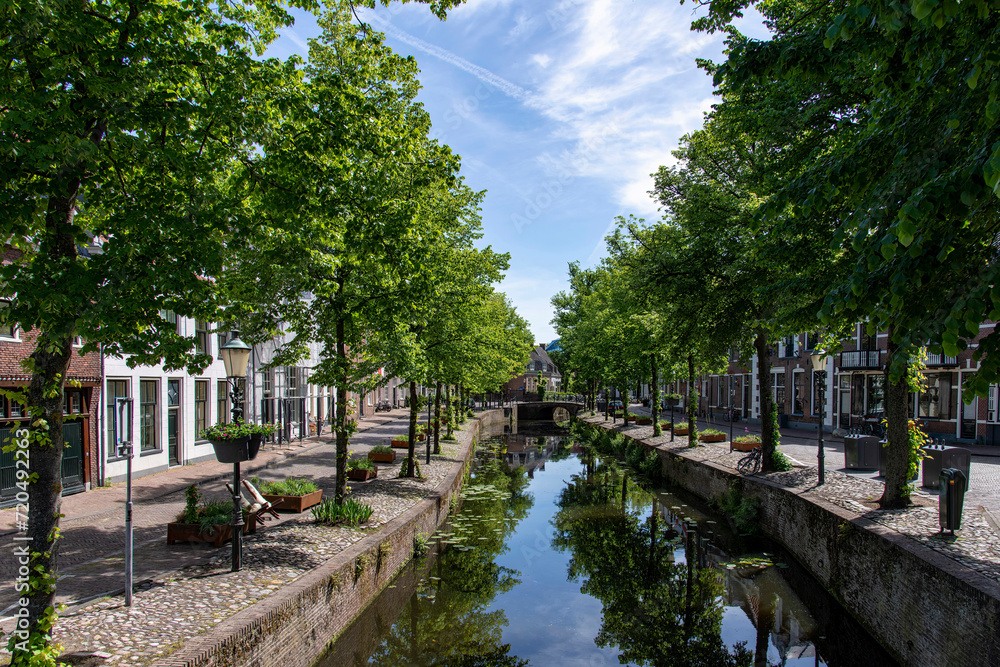View of bridge and street on Havik canal in Amersfoort, the Netherlands, with historic buildings and green trees reflected in the tranquil water of the canal