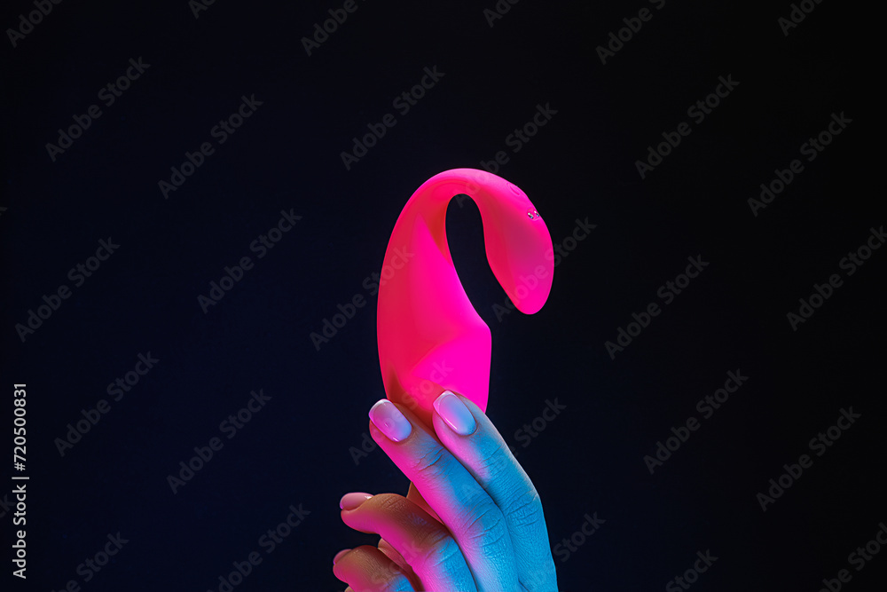 Close-up of a girl holding a red-colored sexual entertainment toy. Sex toy clitoral vibrator on a black background with neon lights. - obrazy, fototapety, plakaty 