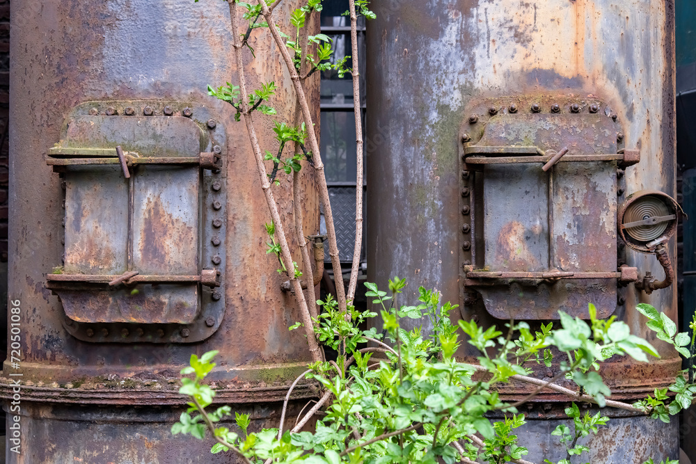 Close up of rusted and weathered furnace or piping system with steel doors of an abandoned and dilapidated industrial steel mill in public Landschaftspark, Duisburg with some green growing in front Close up of rusted and weathered furnace or piping system with steel doors of an abandoned and dilapidated industrial steel mill in public Landschaftspark, Duisburg with some green growing in front