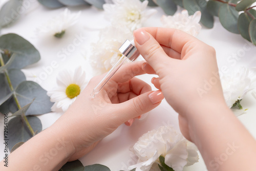 Organic oil for health of nail and skincare concept. Women hands hold pipette with cosmetic serum on white background with flowers, top view
