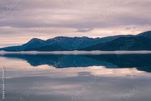 Lake Clark in Lake Clark National Park and Preserve in Alaska. Turquoise lake with rugged mountains, islands and shore. Denaʼina: Qizhjeh Vena. Pink and purple reflections on foggy lake.