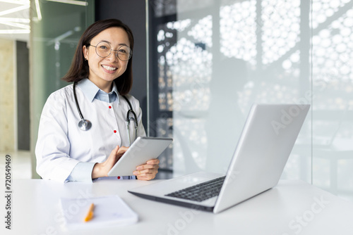 Joyful asian intern woman dressed in medical coat sitting at desktop with laptop and holding digital tablet in hands. Smiling female nurse using modern gadgets for productive work in hospital office.