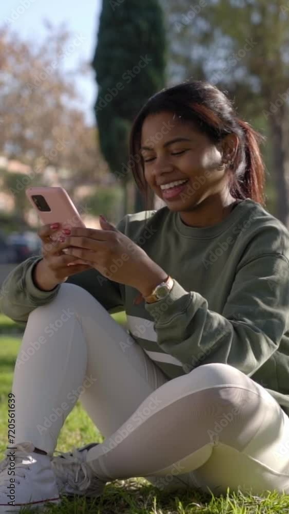 Happy African American woman sitting outdoors on the ground at public park speaking on the phone laughing, spending time in the nature. Vertical video. Happy African American woman sitting outdoors on the ground at public park speaking on the phone laughing, spending time in the nature. Vertical video.