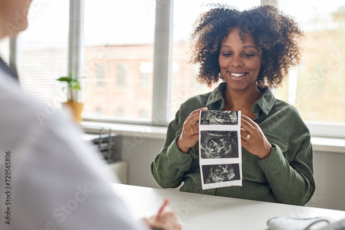 Medium shot of cheerful pregnant Black woman looking at unborn baby sonogram image during consultation in prenatal clinic photo