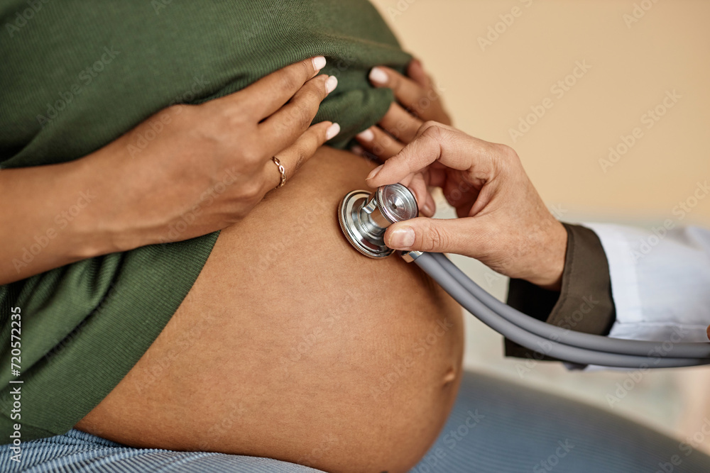 Close up shot of pregnant Black female patients round belly and unrecognizable obstetricians hand conducting auscultation