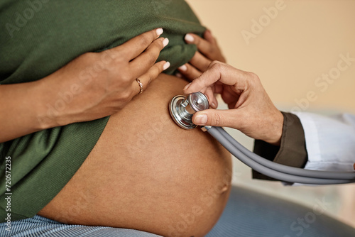 Close up shot of pregnant Black female patients round belly and unrecognizable obstetricians hand conducting auscultation photo