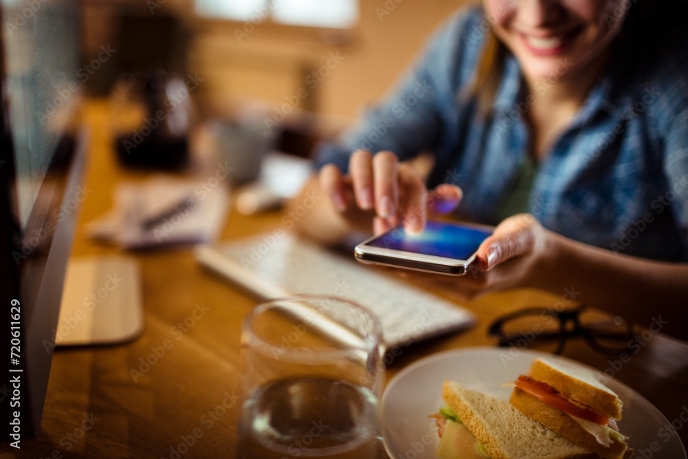 Woman smiling and using smartphone while having breakfast at home computer Woman smiling and using smartphone while having breakfast at home computer