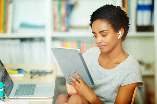 focused black woman uses a tablet with earbuds, effortlessly switching between devices in her productive and tech-savvy home office environment. photo