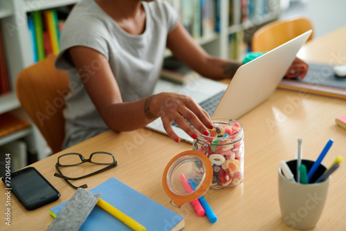 partial view of a woman working from home, reaching for a sweet snack from a jar on her desk filled with colorful candies, next to her laptop and stationery. photo