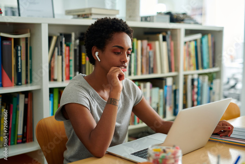 contemplative black young afro woman with earbuds engages deeply with her task on a laptop, her focus uninterrupted in her organized home office setting. photo