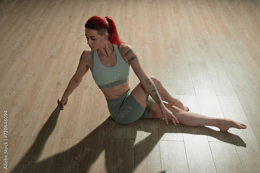 High angle view of tattooed Caucasian girl with red ponytail stretching hips sitting on studio floor in dramatic lighting