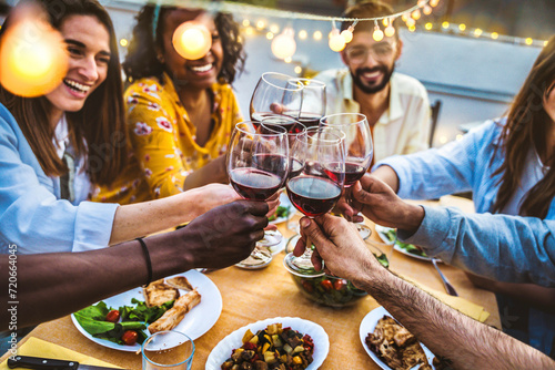 People toasting red wine glasses on rooftop dinner party - Happy friends eating meat and drinking wineglass at restaurant patio - Food and beverage lifestyle concept with guys and girls dining outdoor photo