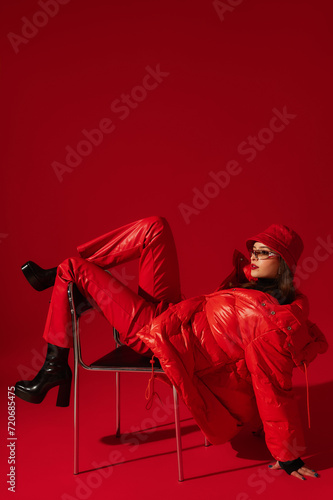 Foto Fashionable confident woman wearing trendy oversized red puffer down jacket, sunglasses, bucket hat, faux leather pants, platform boots, posing on red background