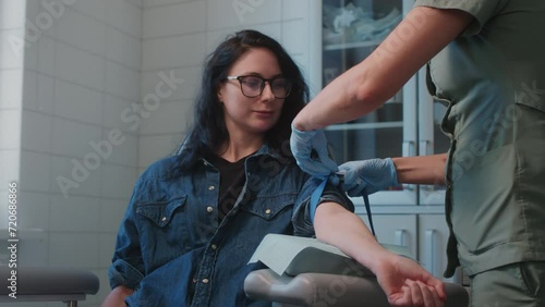 Nurse applies tourniquet and asks woman patient to clench fist for better visibility of vein. Experienced physician explains process to woman before starting blood drawing procedure photo
