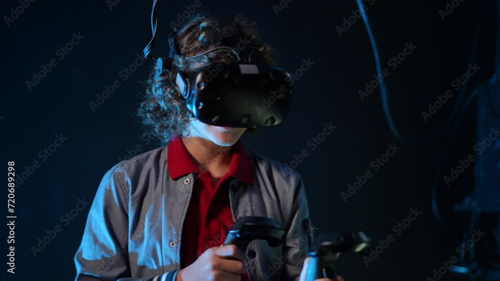 Close-up. In a dark room with blue lighting, a teenager in a VR glasses and with controllers in his hands plays a video game Close-up. In a dark room with blue lighting, a teenager in a VR glasses and with controllers in his hands plays a video game