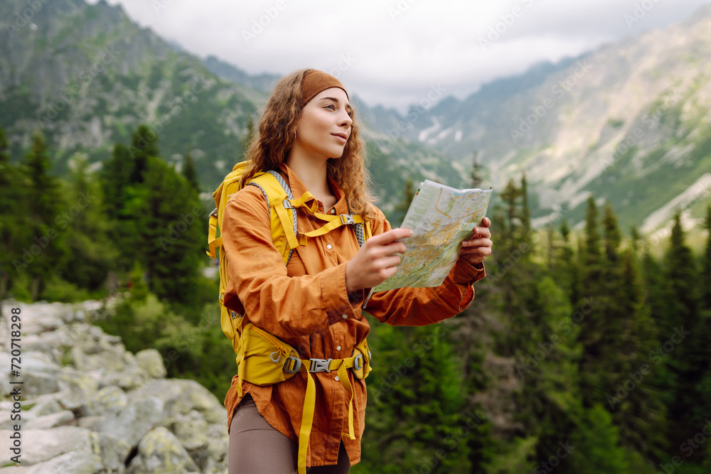 Naklejka premium Young woman traveler with a yellow backpack, holds a map, explores hiking trails in the mountains. The concept of travel, vacation. Hiking and adventure concept