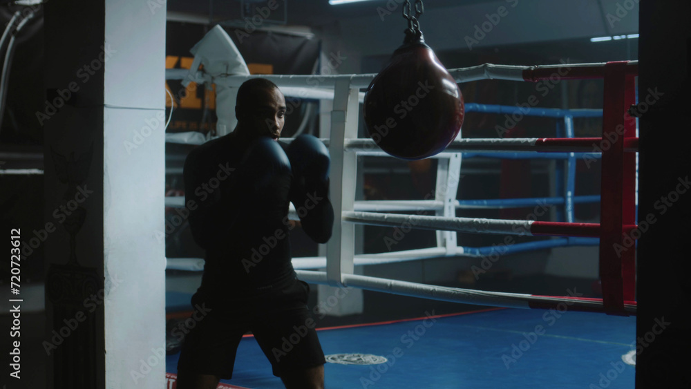 Obraz premium Confident African American fighter hits punching bag while training in dark boxing gym. Athletic man in boxing gloves exercises before fighting competition. Physical activity and intensive workout.