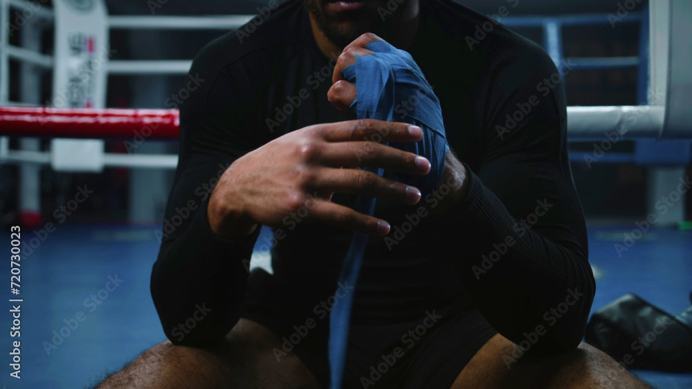 African American boxer sits near boxing ring and wraps his hands with bandage for martial arts. Athlete trains in boxing gym and prepares to fight or competition. Physical activity. Close up. Zoom out