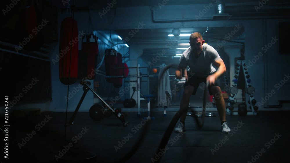 Obraz premium Male athlete exercises with battle ropes in dark boxing gym with LED lighting. Professional boxer does cardio or endurance workout before championship fight. Physical activity and CrossFit training.