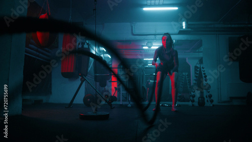 Female athlete exercises with battle ropes in dark boxing gym with LED lighting. Female boxer does cardio or endurance workout before championship fight. Physical activity and CrossFit training.