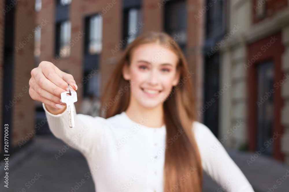 Pretty young woman is standing near  at inner yard of apartment building. She is relocating to a new home and holding a key. She is happy. 