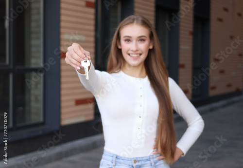 Pretty young woman is standing near at inner yard of apartment building. She is relocating to a new home and holding a key. She is happy. 