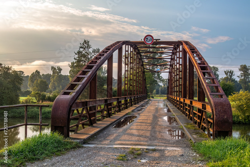 A steel bridge over a water channel connecting dirt roads. There is a sign on the bridge specifying the maximum weight limit for a passing vehicle.