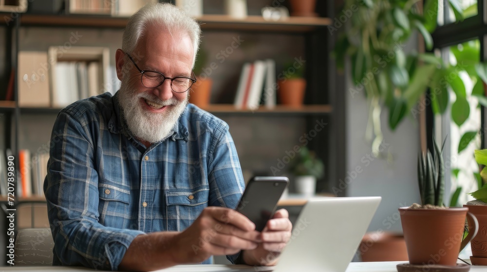 Handsome smiling senior man wearing glasses using mobile phone while sitting at his cozy workplace with laptop at home, retired male chatting with friends in social media Handsome smiling senior man wearing glasses using mobile phone while sitting at his cozy workplace with laptop at home, retired male chatting with friends in social media