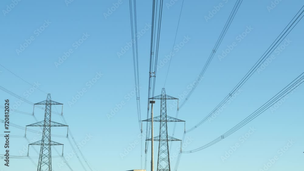 Overhead electric transmission line near a power plant against clear blue sky. Industrial infrastructure with numerous steel towers and wires for distribution of electric energy along large distances. Overhead electric transmission line near a power plant against clear blue sky. Industrial infrastructure with numerous steel towers and wires for distribution of electric energy along large distances.