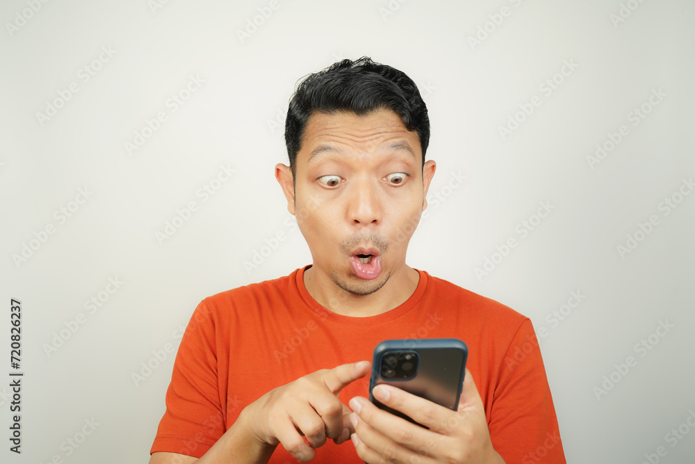 Wow face of Asian man in orange t-shirt shocked what he see in the smartphone on isolated background Wow face of Asian man in orange t-shirt shocked what he see in the smartphone on isolated background
