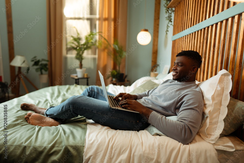 Positive happy African American man, IT specialist using laptop, lying on bed at home, relaxing