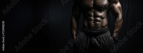 Torso of a muscular man in black pants against a dark background highlighting his abs. © Liana