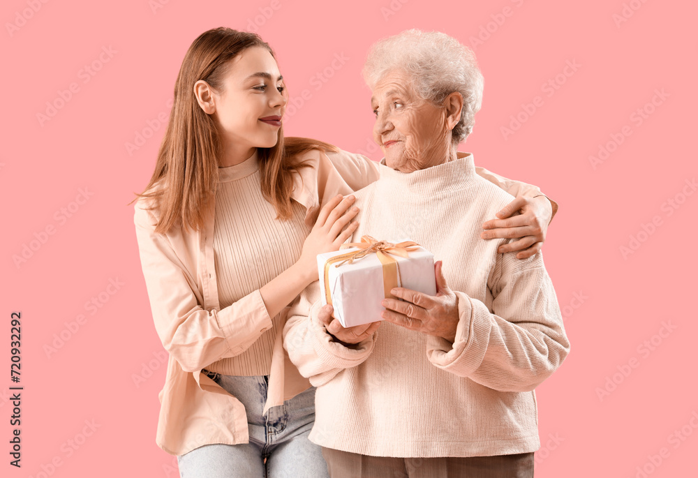 Young woman hugging her grandmother with gift on pink background Young woman hugging her grandmother with gift on pink background