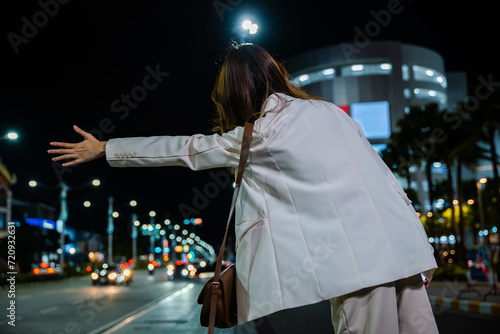 Asian business woman hail waving hand taxi on road in city street at night, Beautiful woman smiling using smartphone application hailing with hand up calling cab outdoor after late work, back view